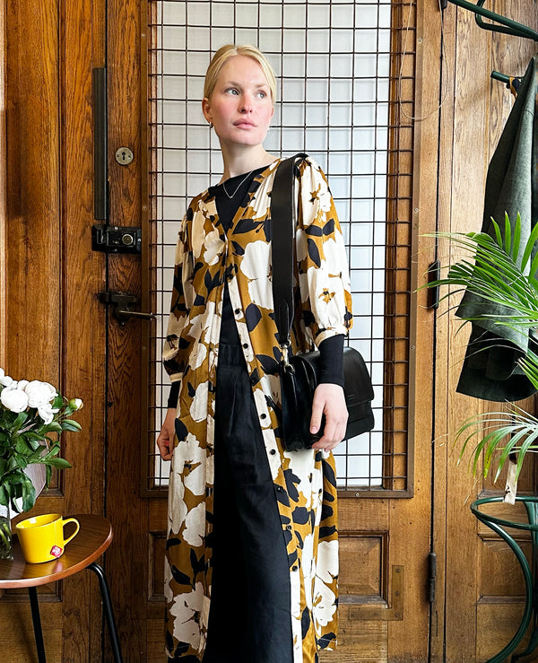 Liebling klänning Bonnie! Bonnie är en smula countryinspirerad i modellen. V-ringad, puffiga ärmar, fickor i sidorna och band att knyta i midjan. Rundat ok bak. Armbågslång ärm med smal manschett med en knapp. Svarta knappar mitt fram. Tyget är blankt vilket ger en lyxig känsla!&nbsp;

Färg: Vita blommor och svarta blad på guldbrun botten.&nbsp;

Material: 51% EcoVero, 49% viskos från Frankrike.
