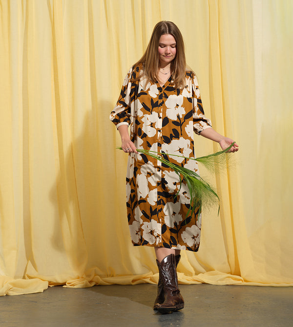 Liebling klänning Bonnie! Bonnie är en smula countryinspirerad i modellen. V-ringad, puffiga ärmar, fickor i sidorna och band att knyta i midjan. Rundat ok bak. Armbågslång ärm med smal manschett med en knapp. Svarta knappar mitt fram. Tyget är blankt vilket ger en lyxig känsla!&nbsp;

Färg: Vita blommor och svarta blad på guldbrun botten.&nbsp;

Material: 51% EcoVero, 49% viskos från Frankrike.