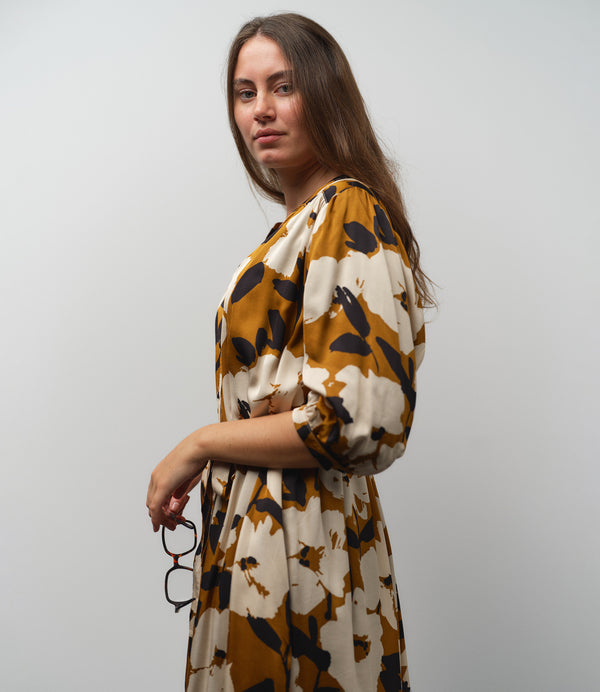 Vår klänning Bonnie! Bonnie är en smula countryinspirerad i modellen. V-ringad, puffiga ärmar, fickor i sidorna och band att knyta i midjan. Rundat ok bak. Armbågslång ärm med smal manschett med en knapp. Svarta knappar mitt fram. Tyget är blankt vilket ger en lyxig känsla!&nbsp;

Färg: Vita blommor och svarta blad på guldbrun botten.&nbsp;

Material: 51% EcoVero, 49% viskos från Frankrike.
Tillverkad i: Litauen
Designad i: Malmö