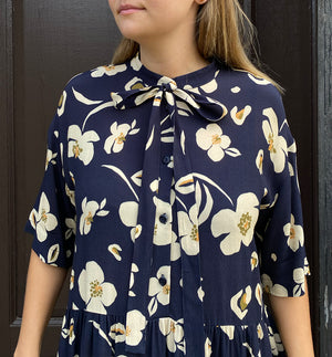  klänning Lyra. Murarkrage med avtagbart band. Knappslå fram med mörkblå knappar. Söm och rynk i midjan. Fickor i sidorna. Bandet knäpps på översta knappen. En fin detalj som ger Lyra fler stylingsmöjligheter. Sydd i en ekologisk viskos. Bandet ingår.

Färg: Gräddvita blommor med gula och olivgröna detaljer på mörkblå botten.

Material:&nbsp;51% EcoVero, 49% viskos från Frankrike. 