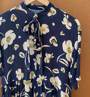  klänning Lyra. Murarkrage med avtagbart band. Knappslå fram med mörkblå knappar. Söm och rynk i midjan. Fickor i sidorna. Bandet knäpps på översta knappen. En fin detalj som ger Lyra fler stylingsmöjligheter. Sydd i en ekologisk viskos. Bandet ingår.

Färg: Gräddvita blommor med gula och olivgröna detaljer på mörkblå botten.

Material:&nbsp;51% EcoVero, 49% viskos från Frankrike. 