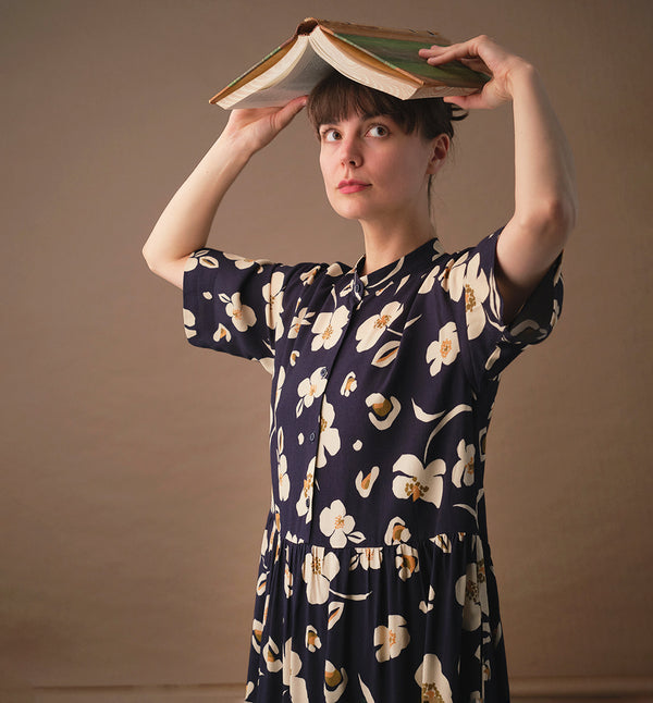 klänning Lyra. Murarkrage med avtagbart band. Knappslå fram med mörkblå knappar. Söm och rynk i midjan. Fickor i sidorna. Bandet knäpps på översta knappen. En fin detalj som ger Lyra fler stylingsmöjligheter. Sydd i en ekologisk viskos. Bandet ingår.

Färg: Gräddvita blommor med gula och olivgröna detaljer på mörkblå botten.

Material:&nbsp;51% EcoVero, 49% viskos från Frankrike. 