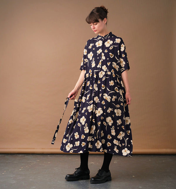 klänning Lyra. Murarkrage med avtagbart band. Knappslå fram med mörkblå knappar. Söm och rynk i midjan. Fickor i sidorna. Bandet knäpps på översta knappen. En fin detalj som ger Lyra fler stylingsmöjligheter. Sydd i en ekologisk viskos. Bandet ingår.

Färg: Gräddvita blommor med gula och olivgröna detaljer på mörkblå botten.

Material:&nbsp;51% EcoVero, 49% viskos från Frankrike. 