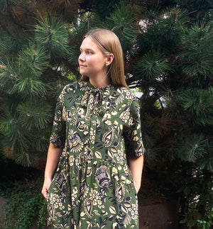 klänning Lyra. Murarkrage med avtagbart band. Knappslå fram med svarta knappar. Söm och rynk i midjan. Fickor i sidorna. Bandet knäpps på översta knappen. En fin detalj som ger Lyra fler stylingsmöjligheter. Sydd i en ekologisk viskos. Bandet ingår.

Färg: Blommor i beige, svart och gulgrönt på grön botten.

Material:&nbsp;51% EcoVero, 49% viskos från Frankrike.