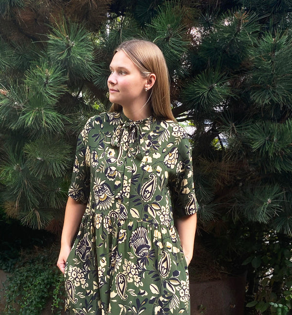 klänning Lyra. Murarkrage med avtagbart band. Knappslå fram med svarta knappar. Söm och rynk i midjan. Fickor i sidorna. Bandet knäpps på översta knappen. En fin detalj som ger Lyra fler stylingsmöjligheter. Sydd i en ekologisk viskos. Bandet ingår.

Färg: Blommor i beige, svart och gulgrönt på grön botten.

Material:&nbsp;51% EcoVero, 49% viskos från Frankrike.