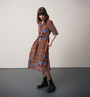 Vår klänning Sally i tyget Tosca.&nbsp;Sally är A-linjeformad med krage, knappslå, ok med små veck i ryggen och fickor i sidorna. Band att knyta i midjan.&nbsp;

Färg: Mönster i kornblått, svart och beige på nougatbrun botten.

Material: 51% EcoVero, 49% viskos från Frankrike.