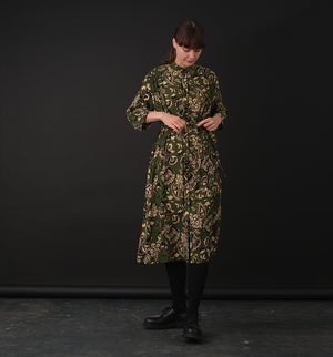 klänning Svala Lotus. En ankellång skjortklänning med lös passform, trekvartsärm och fickor i sidorna. Ok med rynk i ryggen. Ett 2,5 cm brett band i samma tyg medföljer och kan knytas i midjan. Svala är fin både öppen och stängd, med eller utan band. Svarta knappar mitt fram och vid ärmslut. Sydd i en fin viskos.

Svala&nbsp;är generös i storleken,&nbsp;välj gärna en storlek mindre än du brukar.
Se måttabell nedan.

Färg: Blommor i beige, svart och gulgrönt på grön botten.

Material:&nbsp;51% EcoVero, 49% v