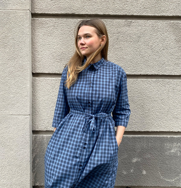 Vår klänning Svala. En ankellång skjortklänning med lös passform, trekvartsärm och fickor i sidorna. Ok med rynk i ryggen. Ett 2,5 cm brett band i samma tyg medföljer och kan knytas i midjan. Svala är fin både öppen och stängd, med eller utan band. Svarta knappar mitt fram och vid ärmslut. Sydd i ett deadstock tyg.&nbsp;

Färg: Svart och blårutig
Material: 100% bomull
Designad i Malmö, sydd i Litauen