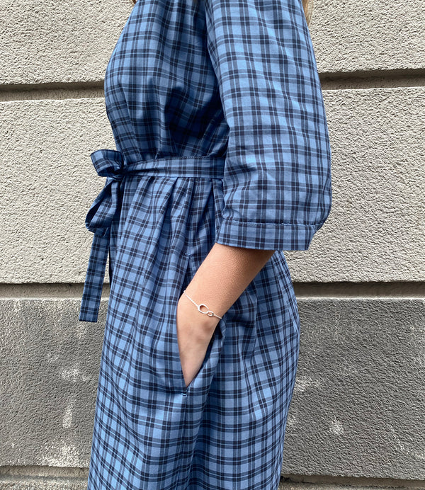 Vår klänning Svala. En ankellång skjortklänning med lös passform, trekvartsärm och fickor i sidorna. Ok med rynk i ryggen. Ett 2,5 cm brett band i samma tyg medföljer och kan knytas i midjan. Svala är fin både öppen och stängd, med eller utan band. Svarta knappar mitt fram och vid ärmslut. Sydd i ett deadstock tyg.&nbsp;

Färg: Svart och blårutig
Material: 100% bomull
Designad i Malmö, sydd i Litauen