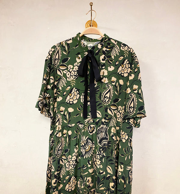 Vår klänning Lyra. Murarkrage med avtagbart band. Knappslå fram med svarta knappar. Söm och rynk i midjan. Fickor i sidorna. Svart band som knäpps på översta knappen. En fin detalj som ger Lyra fler stylingsmöjligheter. Sydd i en ekologisk viskos. Bandet ingår.

Färg: Blommor i beige, svart och gulgrönt på grön botten. Svart band.