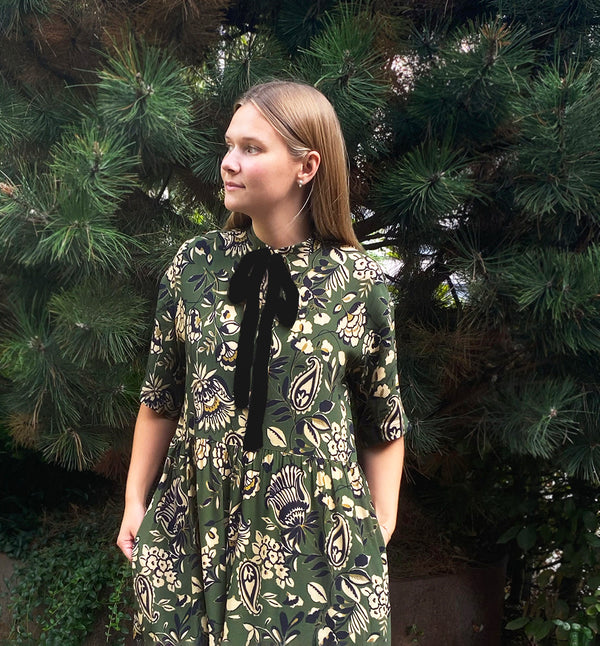 Vår klänning Lyra. Murarkrage med avtagbart band. Knappslå fram med svarta knappar. Söm och rynk i midjan. Fickor i sidorna. Svart band som knäpps på översta knappen. En fin detalj som ger Lyra fler stylingsmöjligheter. Sydd i en ekologisk viskos. Bandet ingår.

Färg: Blommor i beige, svart och gulgrönt på grön botten. Svart band.