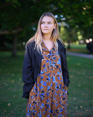 Vår klänning Sally i tyget Tosca.&nbsp;Sally är A-linjeformad med krage, knappslå, ok med små veck i ryggen och fickor i sidorna. Band att knyta i midjan.&nbsp;

Färg: Mönster i kornblått, svart och beige på nougatbrun botten.

Material: 51% EcoVero, 49% viskos från Frankrike.